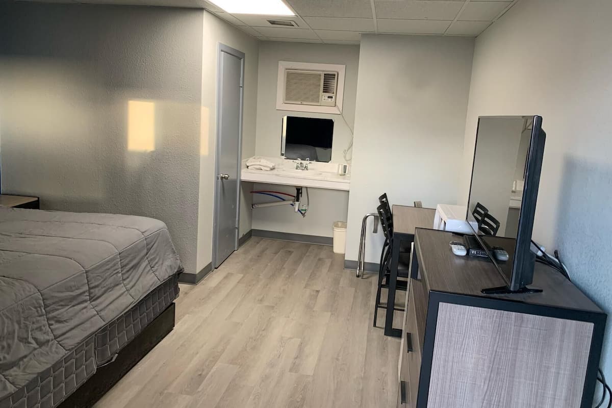 A hotel room with a bed, a desk with a computer, a mirror, a window, and a bathroom with a sink and toilet.