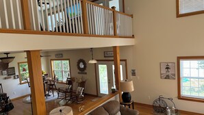 A spacious living area with a high ceiling, wooden railing, and a view of the outdoors through large windows. - Cozy River View House (Nanjemoy)