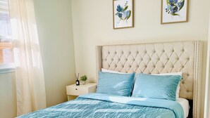 Ein Schlafzimmer mit einer gepolsterten Kopfteile, einem Bett mit blauen, gemusterten Bettdecken, zwei gerahmten floralen Kunstwerken an der Wand und einem Nachttisch mit einer kleinen Pflanze.