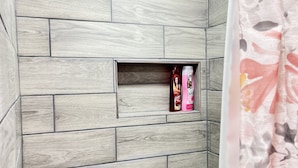 A bathroom with a shower area featuring a wooden wall design and a small shelf with toiletries. - Private Suite with Washer and Dryer, Kitchenette  and Enclosed Parking (Las Cruces)