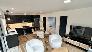 Ein modernes Wohnzimmer mit einem grauen Sofa, einem runden Couchtisch und zwei weißen Sessel. An einer schwarzen Unterhaltungsecke ist ein Flachbildfernseher angebracht. Der Raum verfügt über eine Küche mit schwarzen Schränken und einer Mikrowelle.