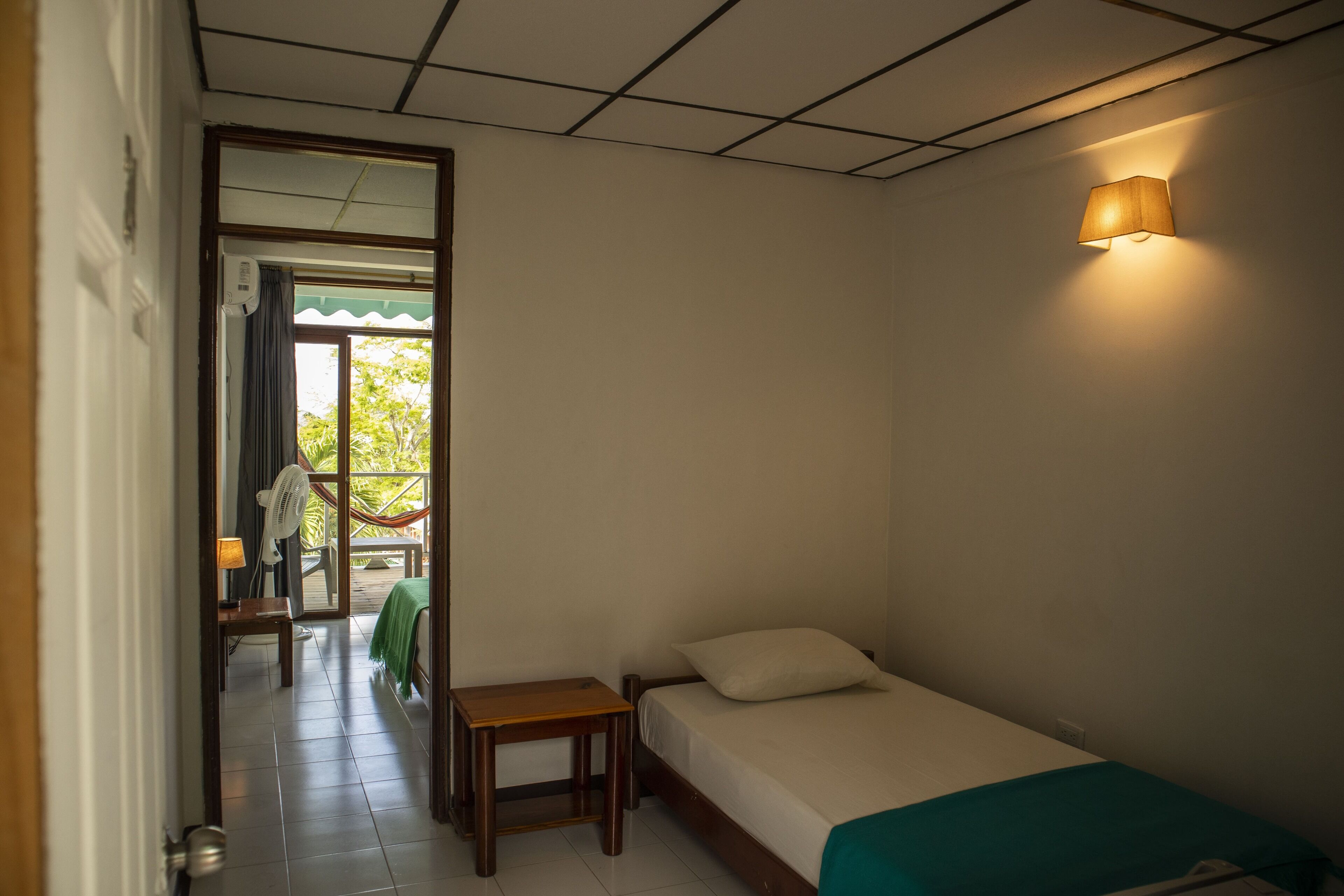 Una habitación pequeña con cama individual, cabecera de madera, mesita de noche, ventilador y ventana con vistas a zonas verdes.