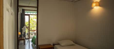Una habitación pequeña con cama individual, cabecera de madera, mesita de noche, ventilador y ventana con vistas a zonas verdes.