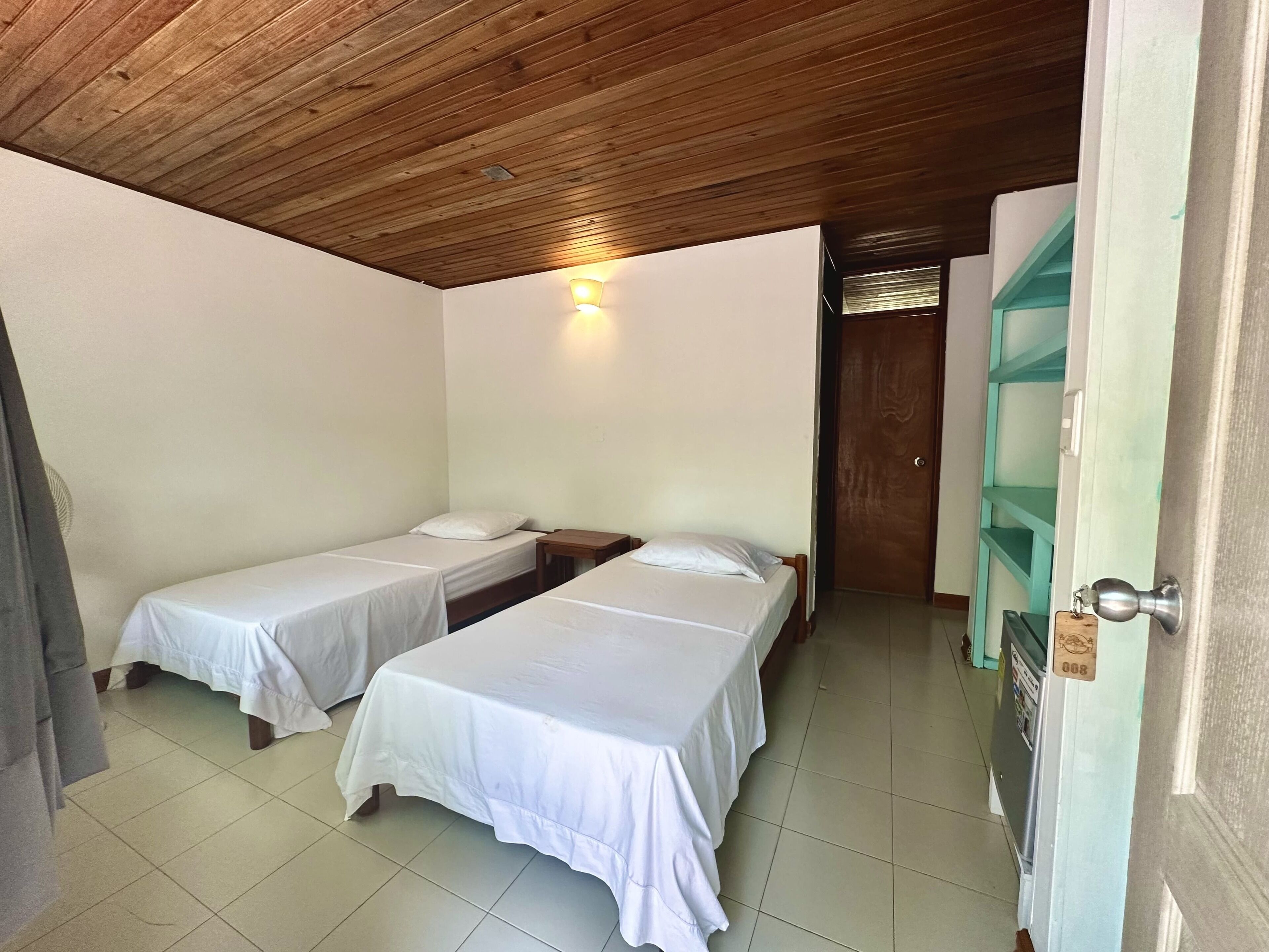 A room with two single beds, a wooden ceiling, a wall-mounted light, and a door with a key lock.