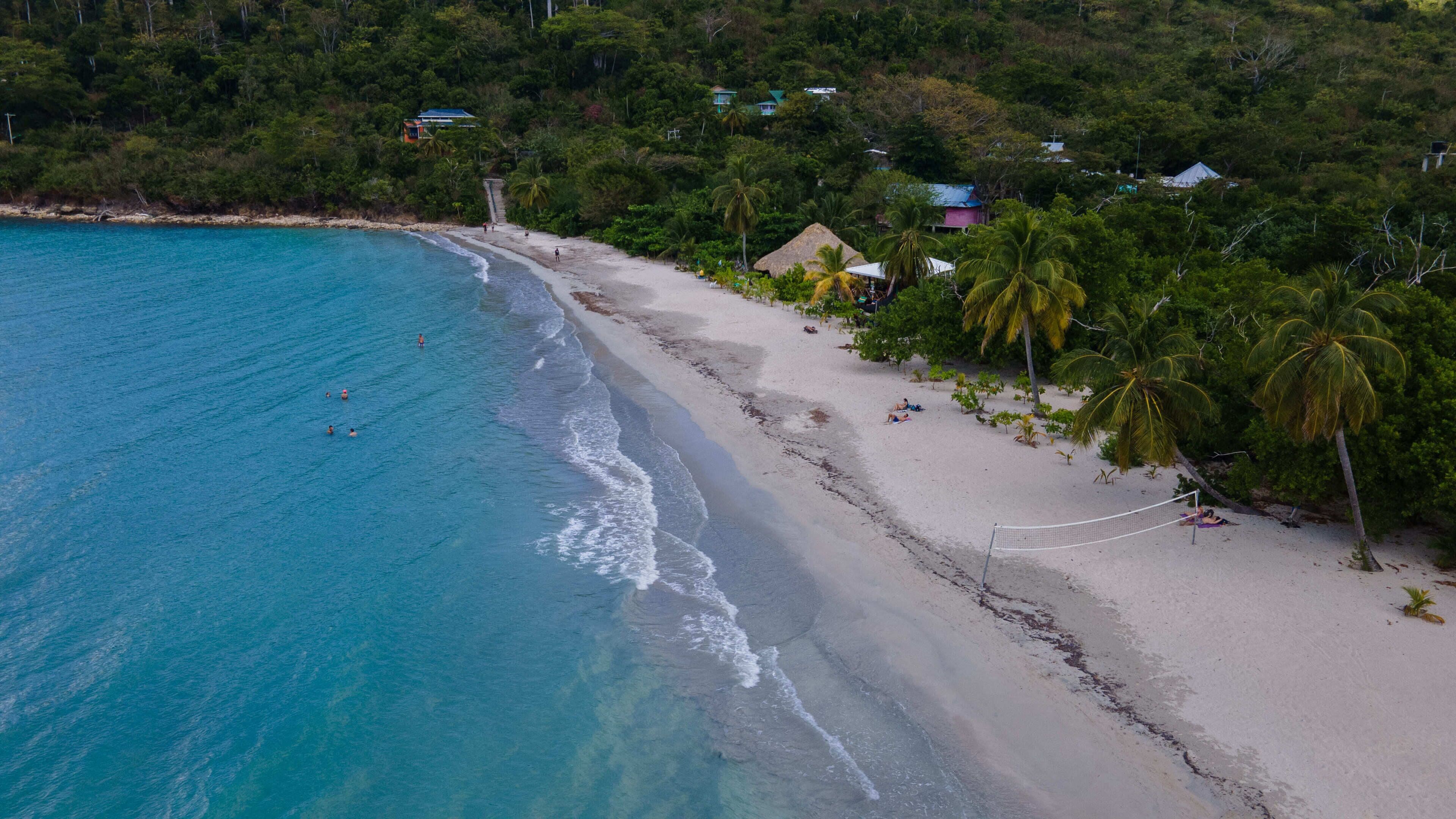 An aerial view of a beach with clear turquoise waters, a sandy shore, and several buildings with thatched roofs among lush greenery.