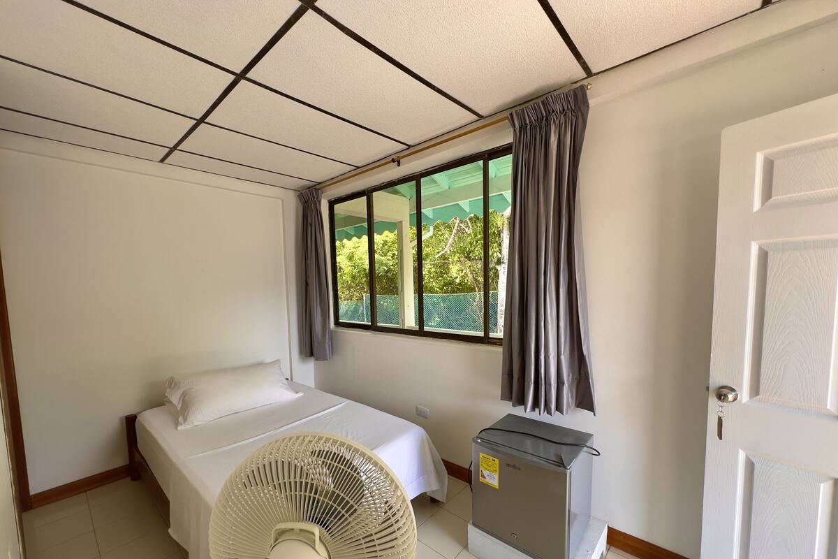 Une chambre d’hôtel avec un lit, un ventilateur sur pied, un radiateur et une fenêtre avec des rideaux.