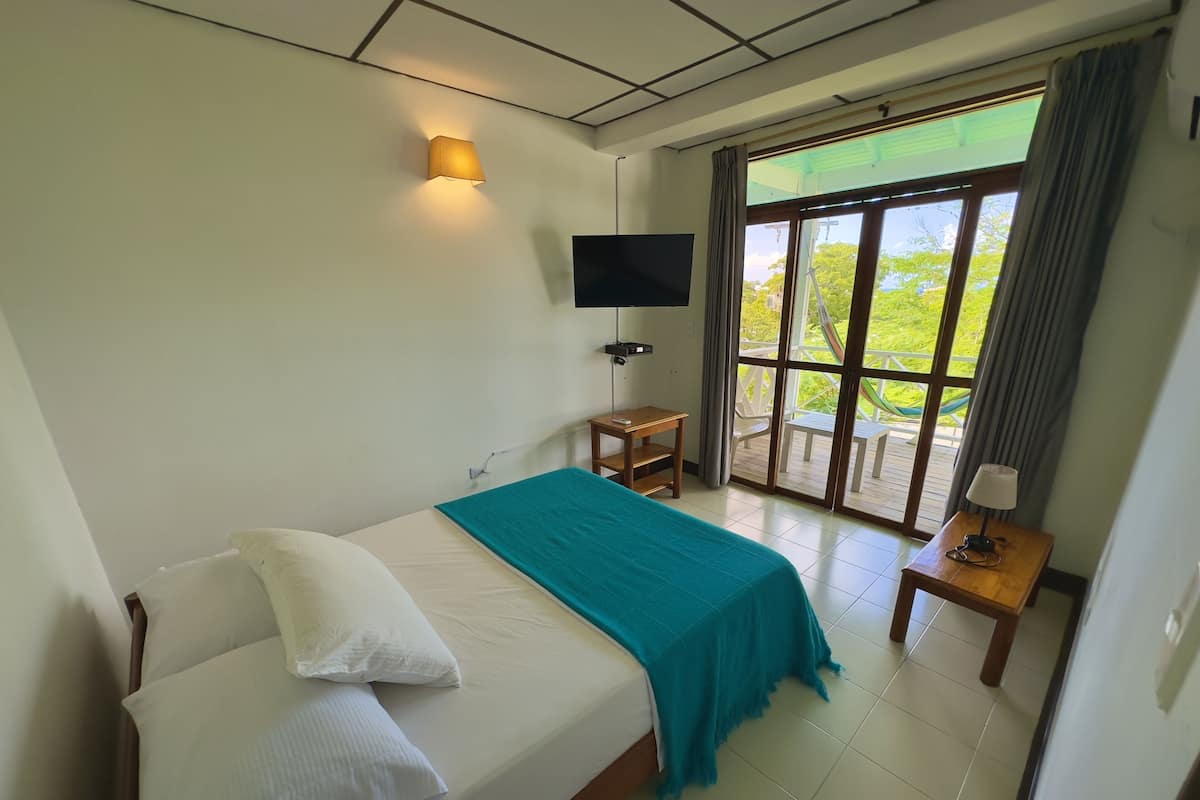 Une chambre d’hôtel comprenant un lit, un téléviseur fixé au mur, une petite table avec une lampe et une porte-fenêtre coulissante donnant sur un espace extérieur.