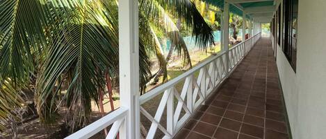 A covered corridor with a tiled floor, white railings, and palm trees.