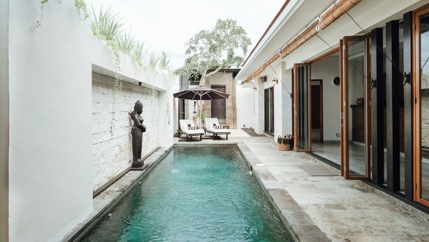 Una piscina con dos sillas de descanso y una estatua, rodeada por un muro blanco y un techo cubierto de bambú.