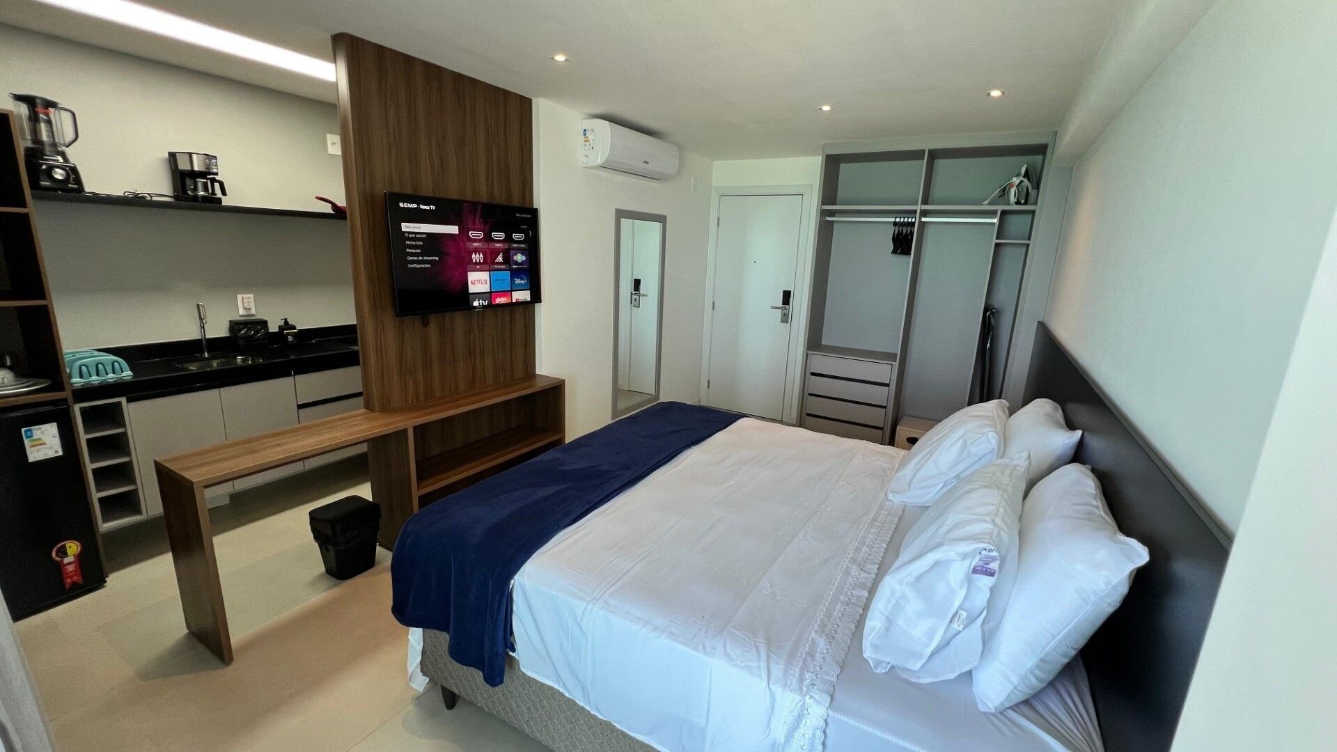 A hotel room with a bed, a TV, a wardrobe, a bench, and a kitchenette.