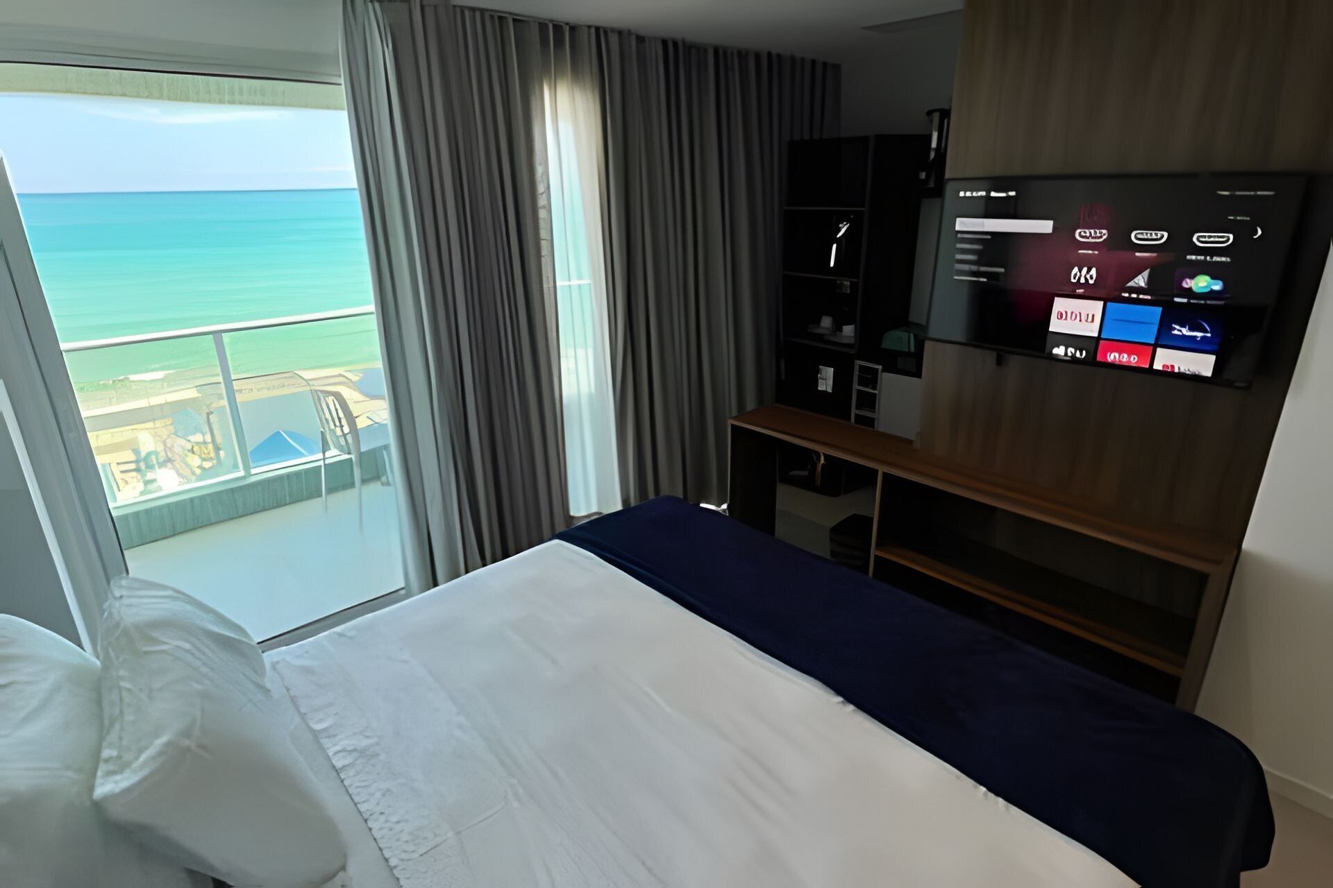 A hotel room with a bed, a TV, a desk, and a view of the ocean.