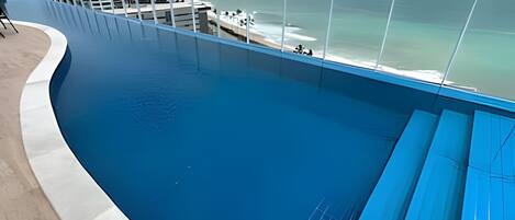 Una grande piscina a forma di curva con acqua cristallina e blu, circondata da un solarium bianco e ringhiere in vetro.