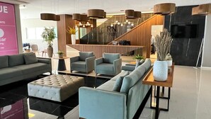 A modern lobby with sofas, armchairs, and a coffee table. There is a staircase with wooden railings and a black wall with a TV screen.