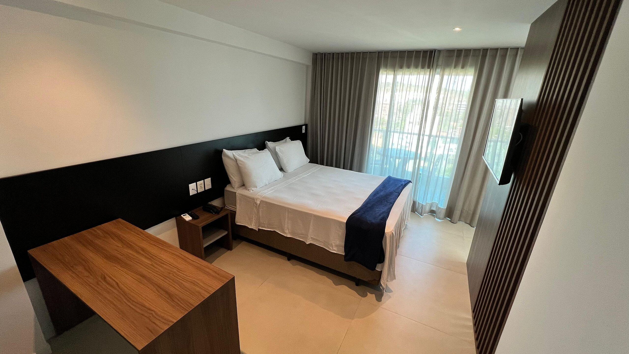 A hotel room with a bed, bedside table, and a TV mounted on the wall.