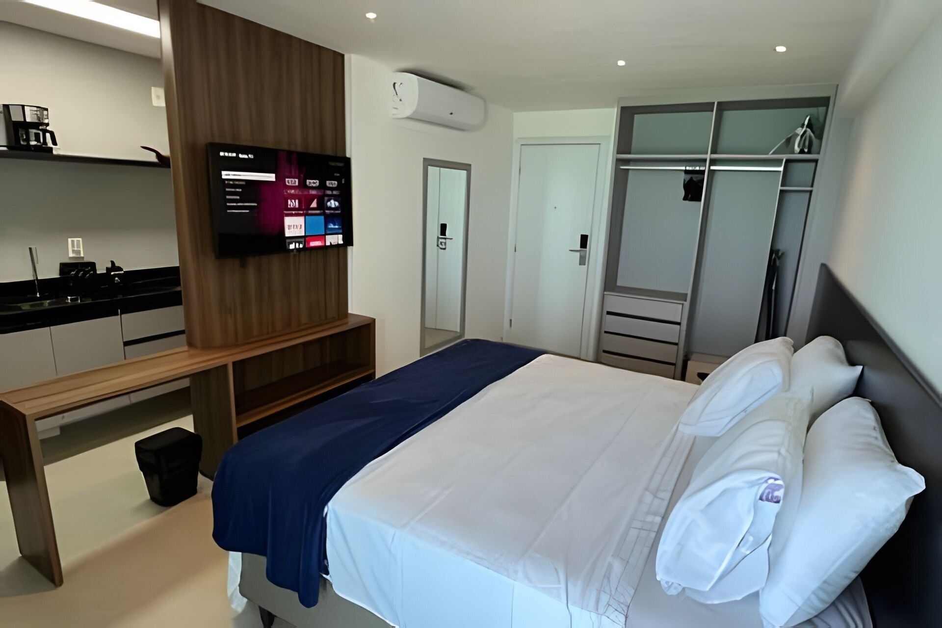 A modern hotel room with a bed, a TV mounted on the wall, a wardrobe, and a small desk with a chair.