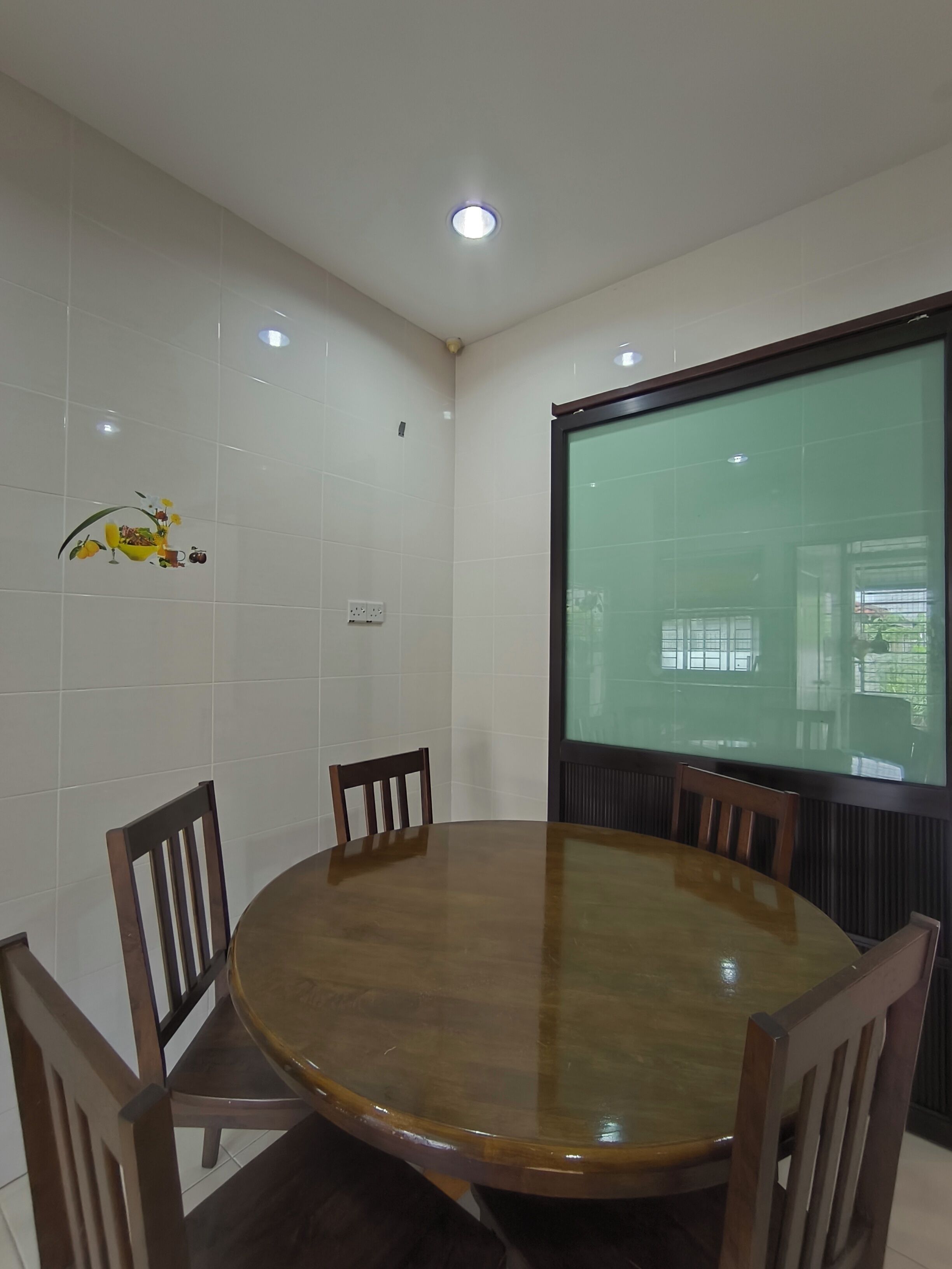 Un salón de conferencias con una mesa redonda, cuatro sillas de madera, paredes de baldosas blancas, una lámpara en el techo y un ventanal con vistas a la vegetación.