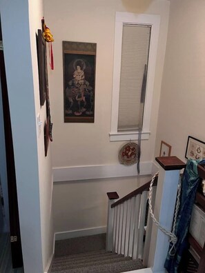 Een trap met een leuning, een aan de muur hangend schilderij en een raam met jaloezieën.