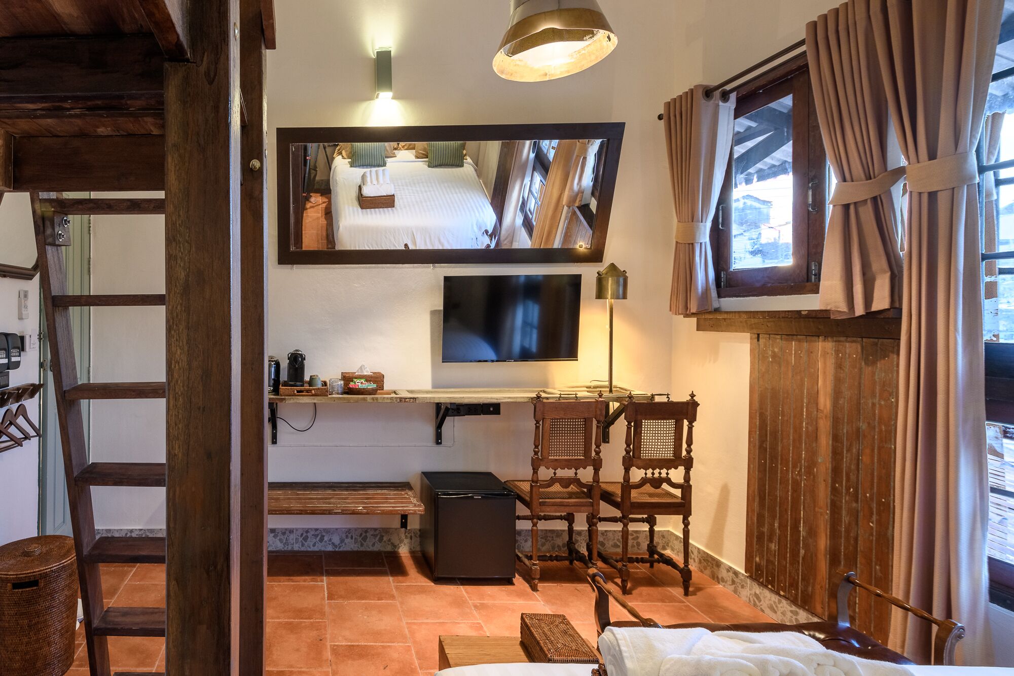 A cozy room with a bed, a television, a desk with a lamp, and a window with curtains.
