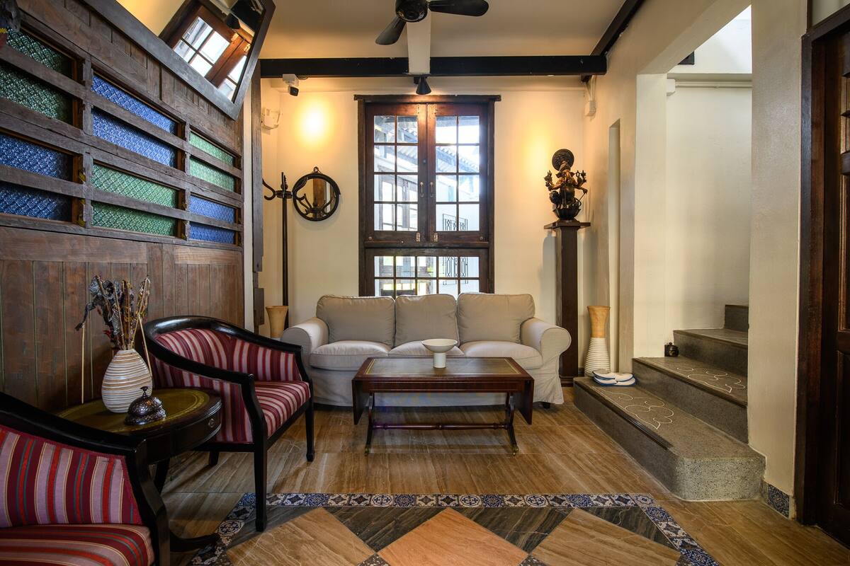A cozy living room with a sofa, armchair, and coffee table. There is a wooden wall with glass panels, a fireplace, and a staircase.