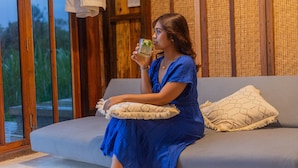 A person sitting on a grey sofa with a cushion, holding a glass of drink, in a room with wooden walls and a window. - Calidas Hill (Kintamani)