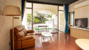 A modern living room with a leather sofa, a glass coffee table, a TV, and a sliding glass door leading to an outdoor area with a garden view. - Higashi Kumamoto Business Center (Ozu)
