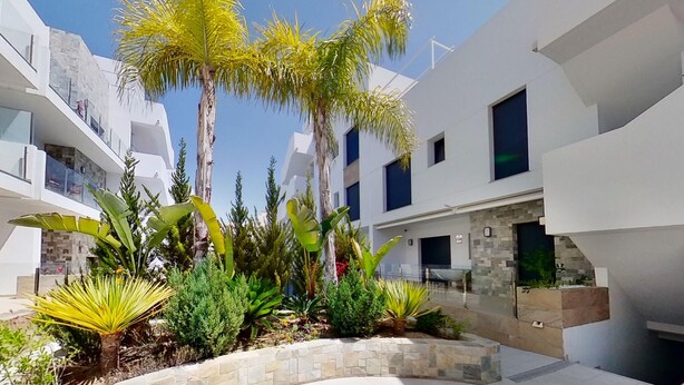 A modern building with white exterior walls, multiple balconies, and a landscaped garden with various plants.