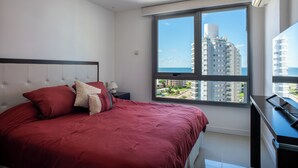 A bedroom with a large bed, a nightstand, a desk, and a window with a view of a high-rise building and the sea. - Departamento en Punta del Este (Punta del Este)