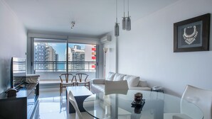 A modern living room with a glass table, white sofas, and a city view through large windows. - Departamento en Punta del Este (Punta del Este)