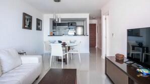 A modern living room with a white sofa, a dining table with chairs, a TV, and a kitchen area with a refrigerator, oven, and microwave. - Departamento en Punta del Este (Punta del Este)