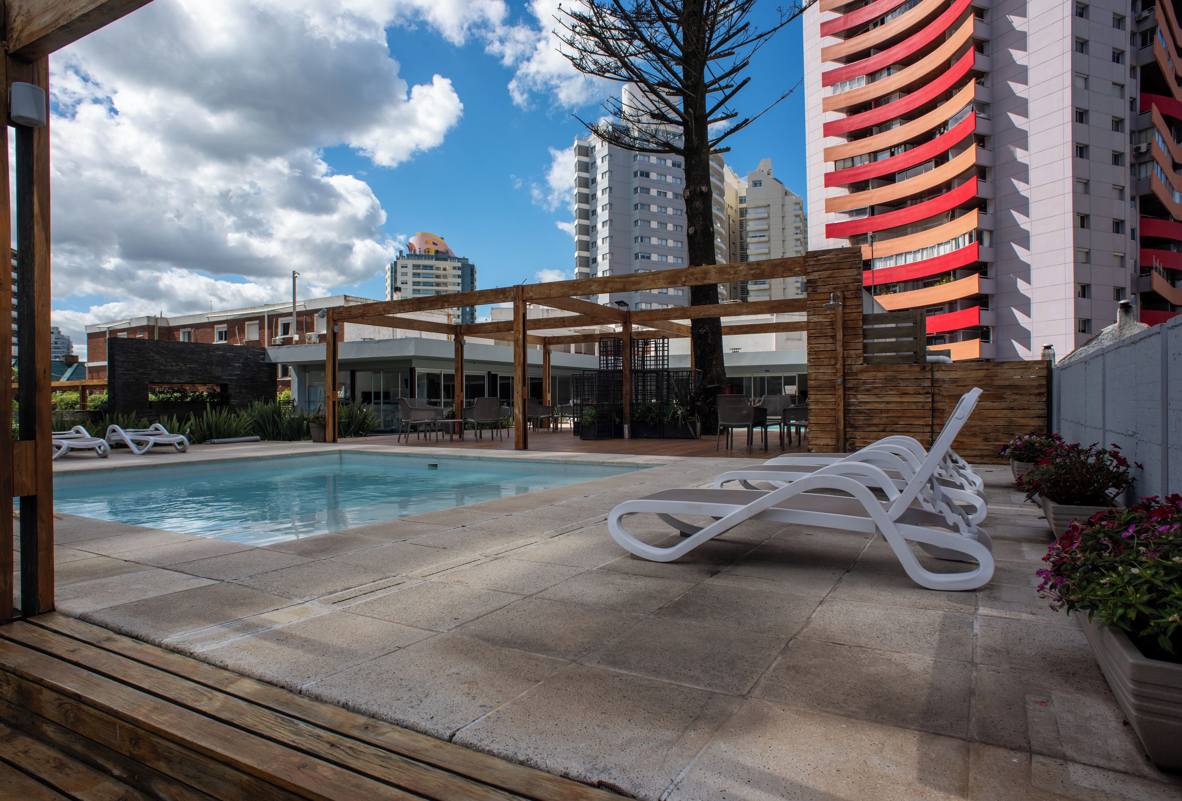 A pool area with lounge chairs and a pergola, surrounded by tall buildings and a clear sky.