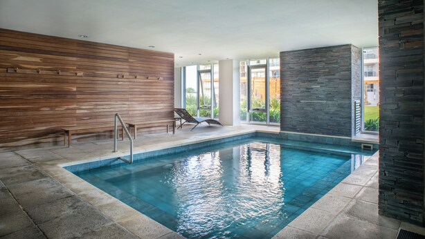 An indoor swimming pool with wooden and stone walls, a clear view of the outside, and a sun lounger by the pool. - Departamento en Punta del Este (Punta del Este)