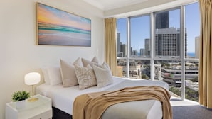 A hotel room with a large bed, a nightstand, a lamp, a painting, and a view of the city. - Chevron Renaissance Ocean View (Surfers Paradise)