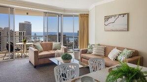 A modern living room with a large window overlooking a cityscape and the sea. There are two sofas, a dining table, and a small balcony area with a table and chairs. - Chevron Renaissance Ocean View (Surfers Paradise)