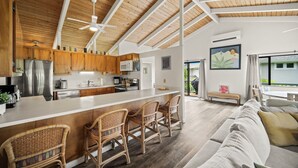 A spacious kitchen with wooden cabinets, a central island with bar stools, a ceiling fan, and a large window overlooking a balcony. - Paradise at Prettyville Kauai, waterfall & ocean View, AC, sleep 8-10, BigAloha (Princeville)