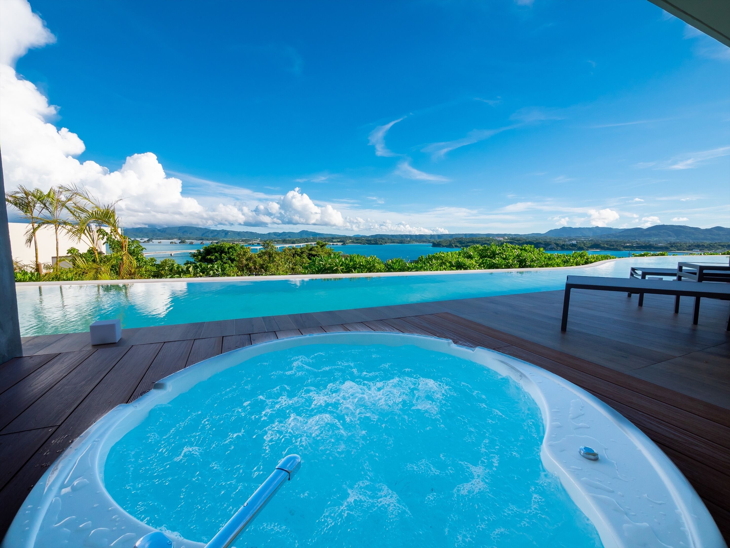 A hot tub with a wooden deck, overlooking a pool and a scenic view of the ocean and mountains.
