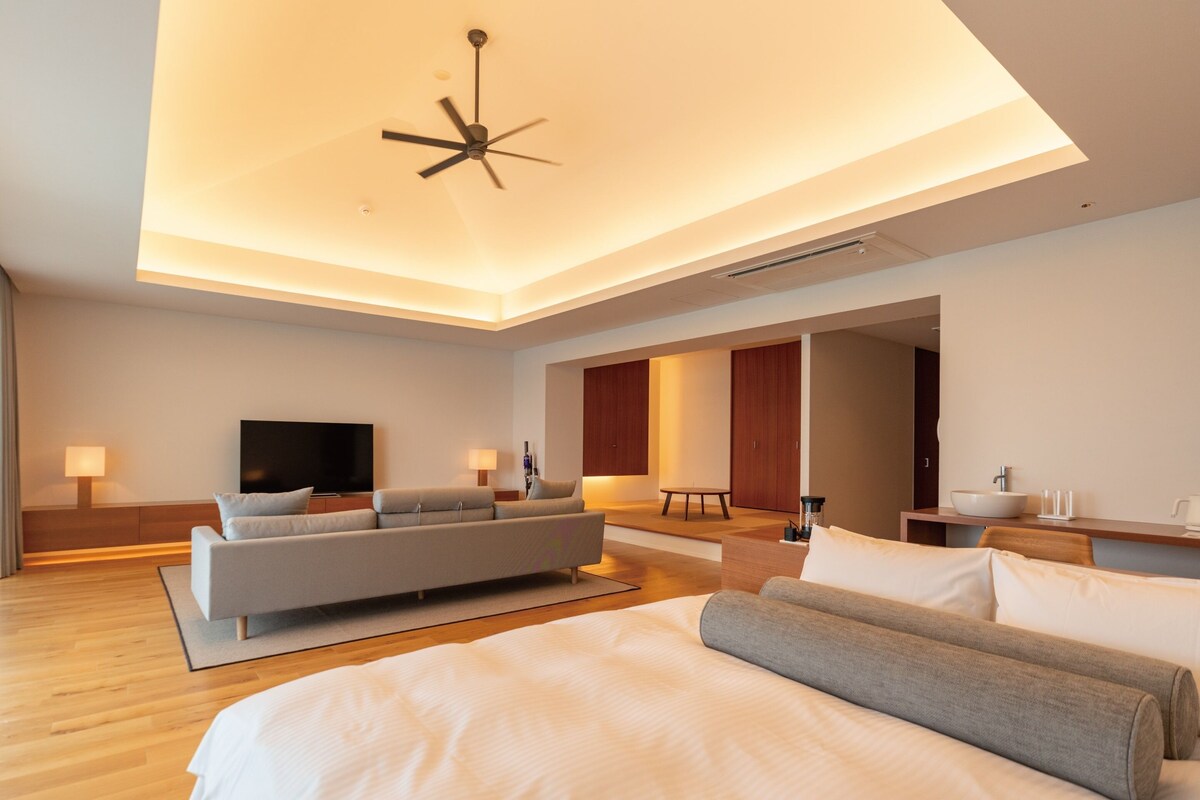 A modern hotel room with a large bed, a sofa, a TV, and a ceiling fan.