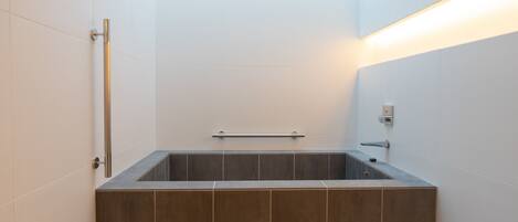A modern bathroom with a rectangular bathtub, a handrail, and a wall-mounted light fixture.