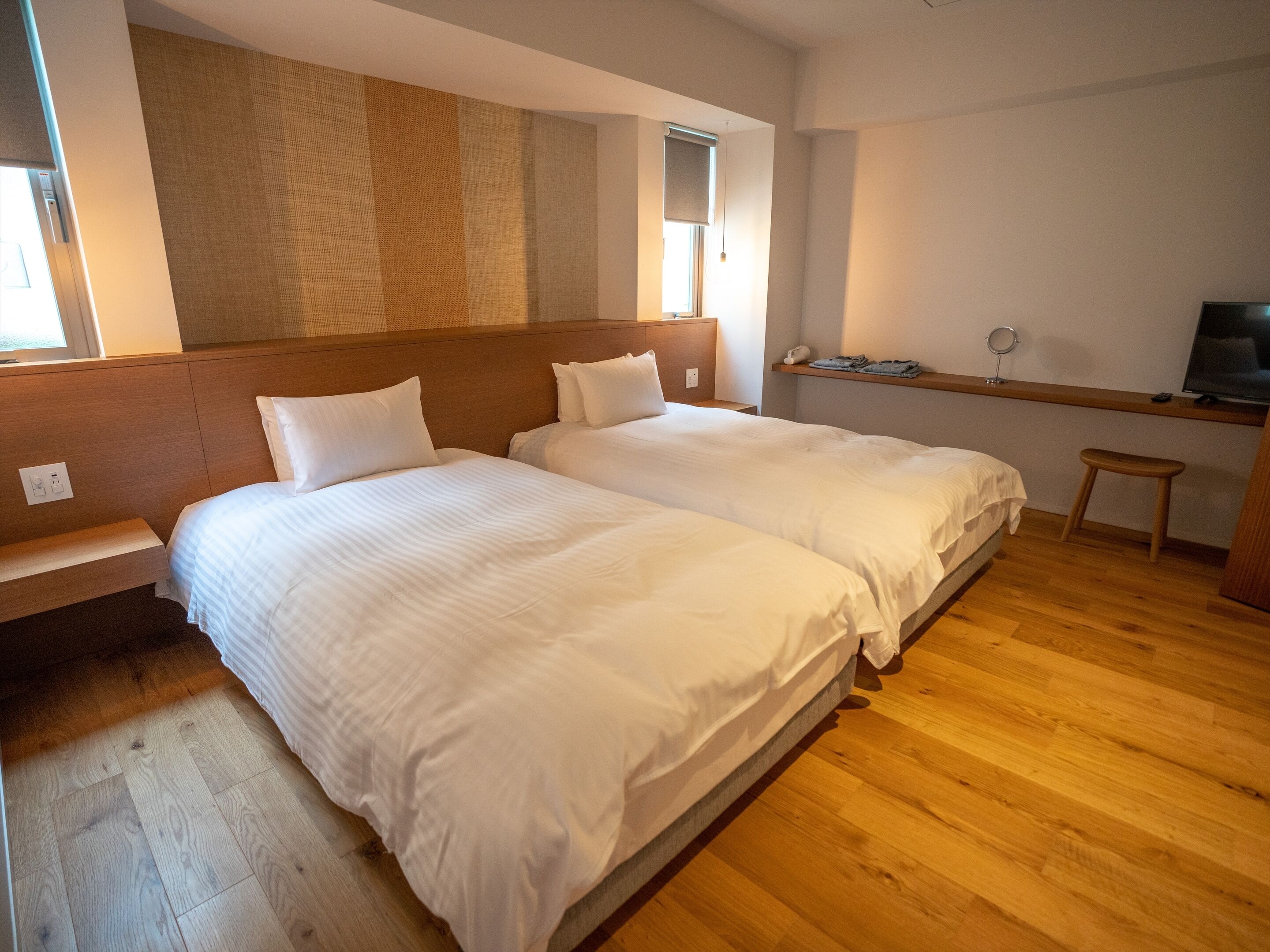 Une chambre d’hôtel avec deux lits, une tête de lit en bois, un petit tabouret, une télévision et une fenêtre avec des stores.