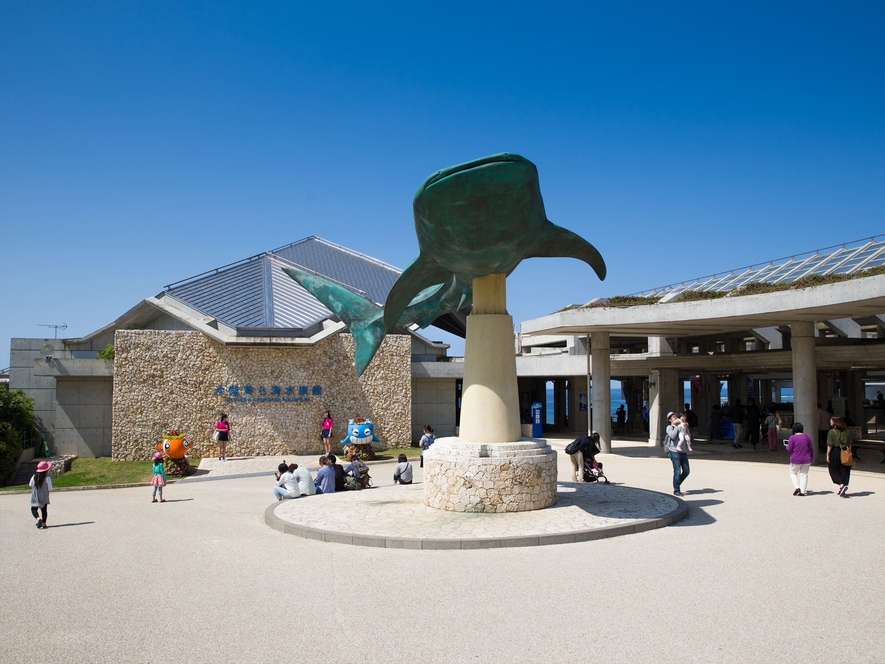 A large sculpture of a fish in front of a building with a stone facade and a blue sign.