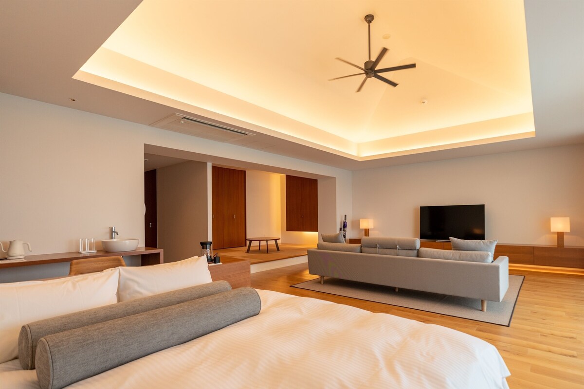 A modern hotel room with a large bed, a sofa, a TV, and a ceiling fan.