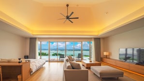 A spacious hotel room with a large bed, a sofa, a TV, and a balcony with a view of the sea and mountains.