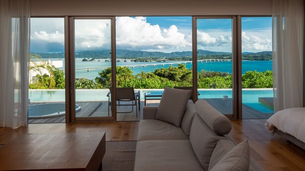A modern living room with a large sliding glass door leading to an infinity pool overlooking a scenic coastal view.