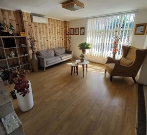Ein Wohnzimmer mit einer Couch, einem Sessel und einem kleinen Tisch mit einer Pflanze. Ein Bücherregal und ein großes Fenster mit Jalousien sind ebenfalls vorhanden.
