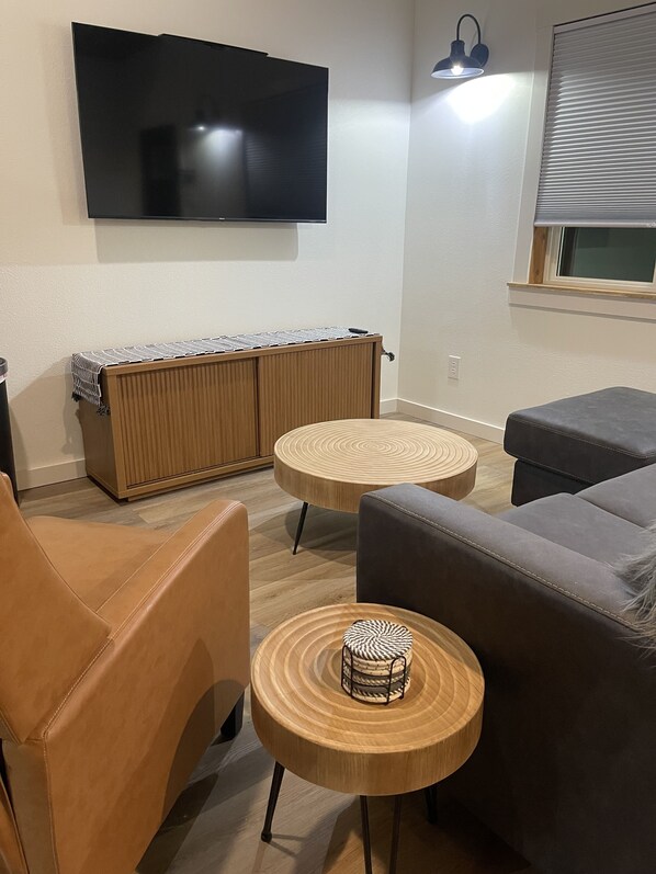 A modern living room with a flat-screen TV mounted on the wall, a wooden radiator, two round wooden tables, a leather armchair, and a grey sofa.