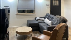 Una sala de estar acogedora con techo de madera, un sofá gris, un sillón de cuero marrón, una mesa de centro redonda de madera y una puerta negra.