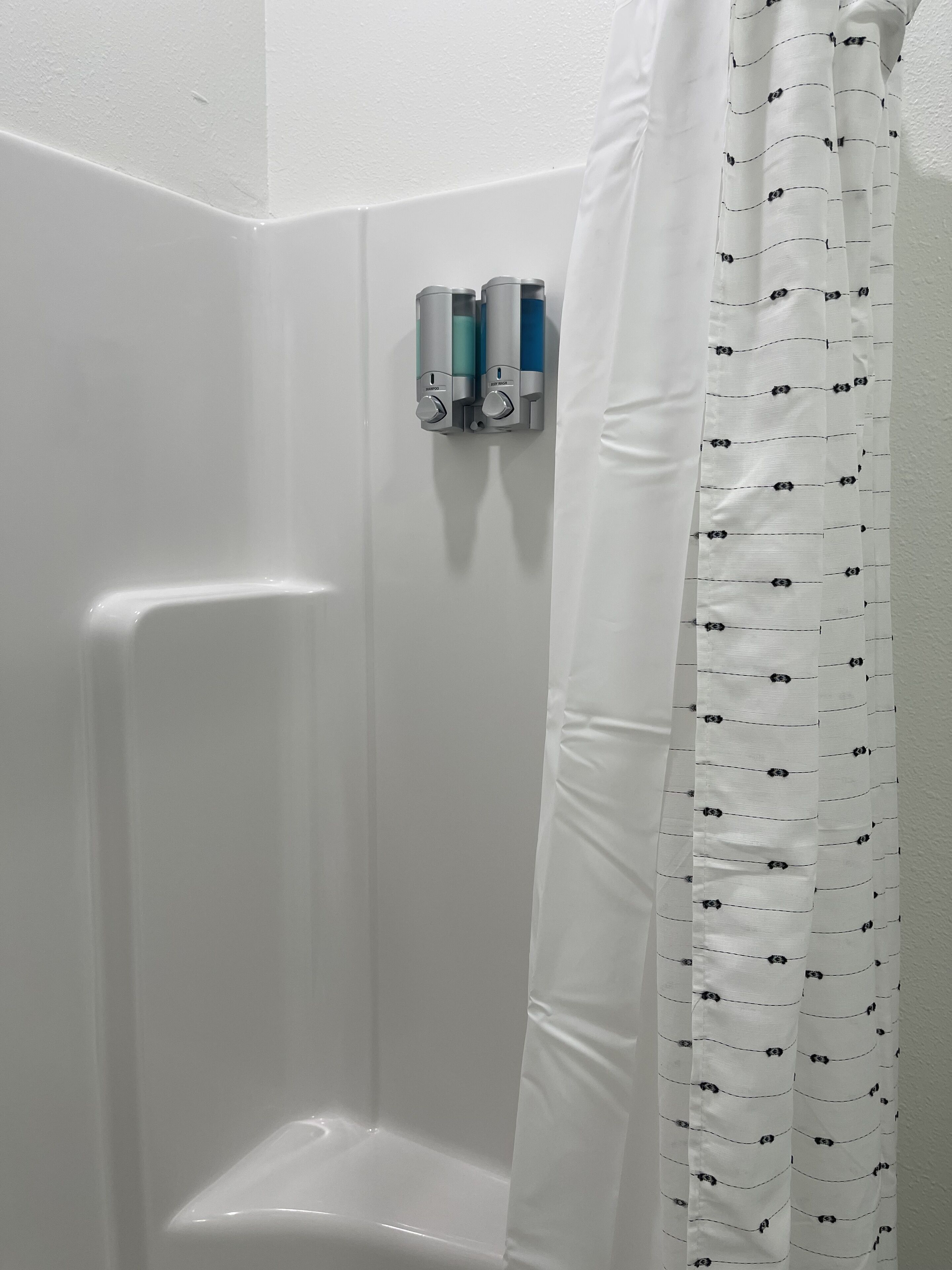 Un angolo di un bagno con una tenda da doccia bianca e un dispenser di sapone fissato al muro.