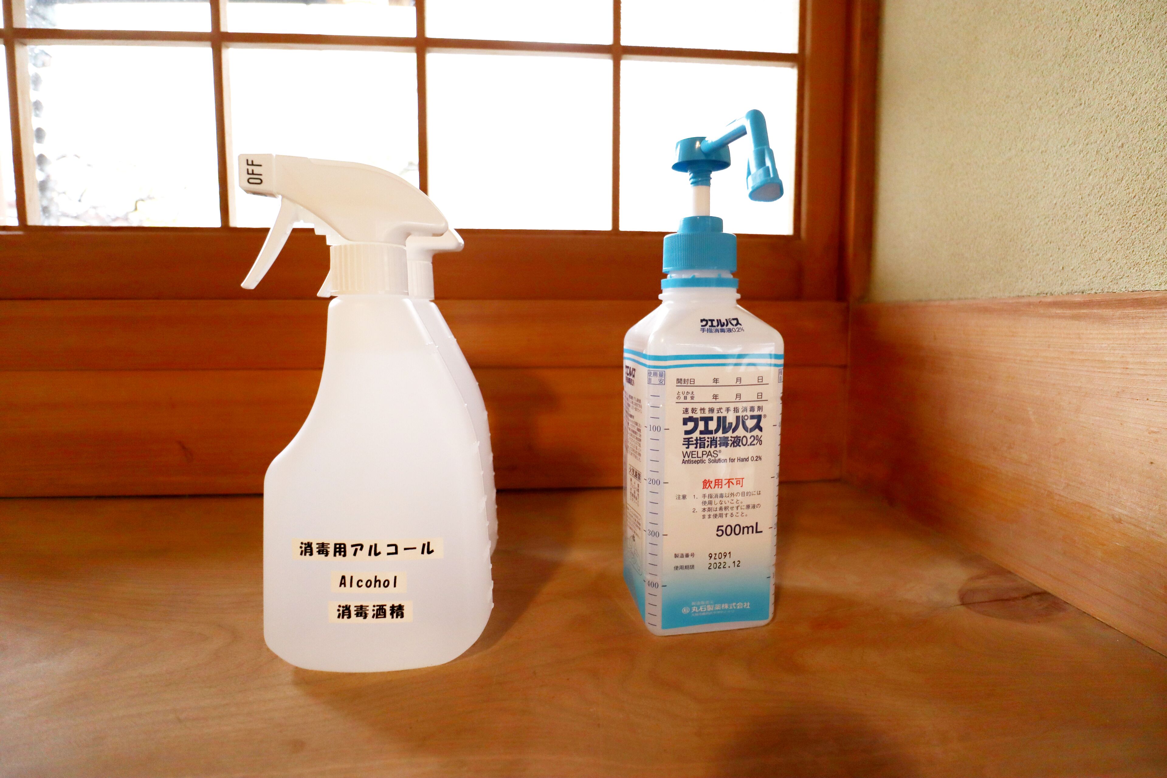 Een sprayfles en een fles WELPAS handdesinfecter op een houten oppervlak nabij een raam.