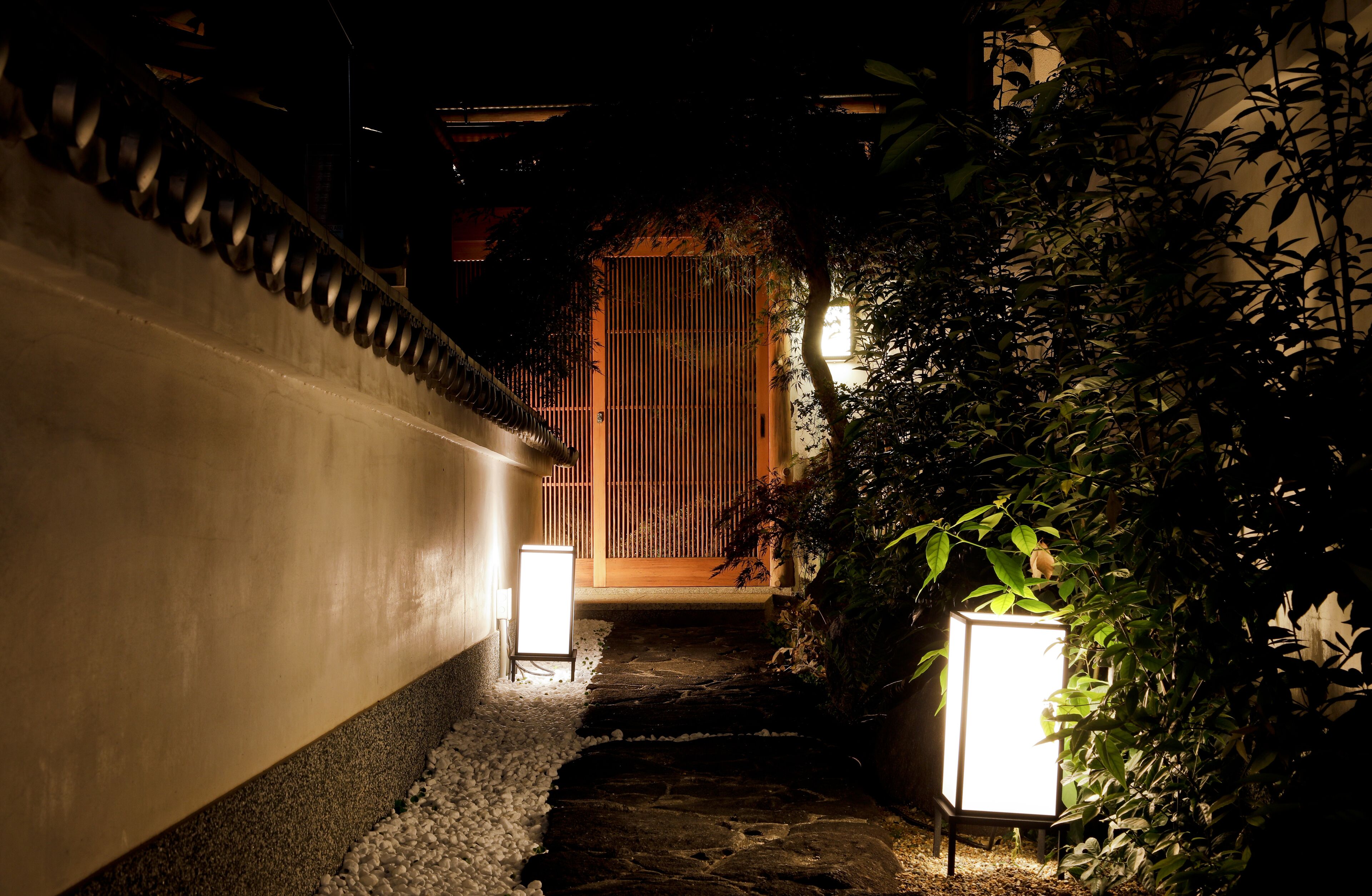 Um caminho de pedra estreito conduz a um portão de madeira tradicional, ladeado por paredes brancas e iluminado por duas lanternas.