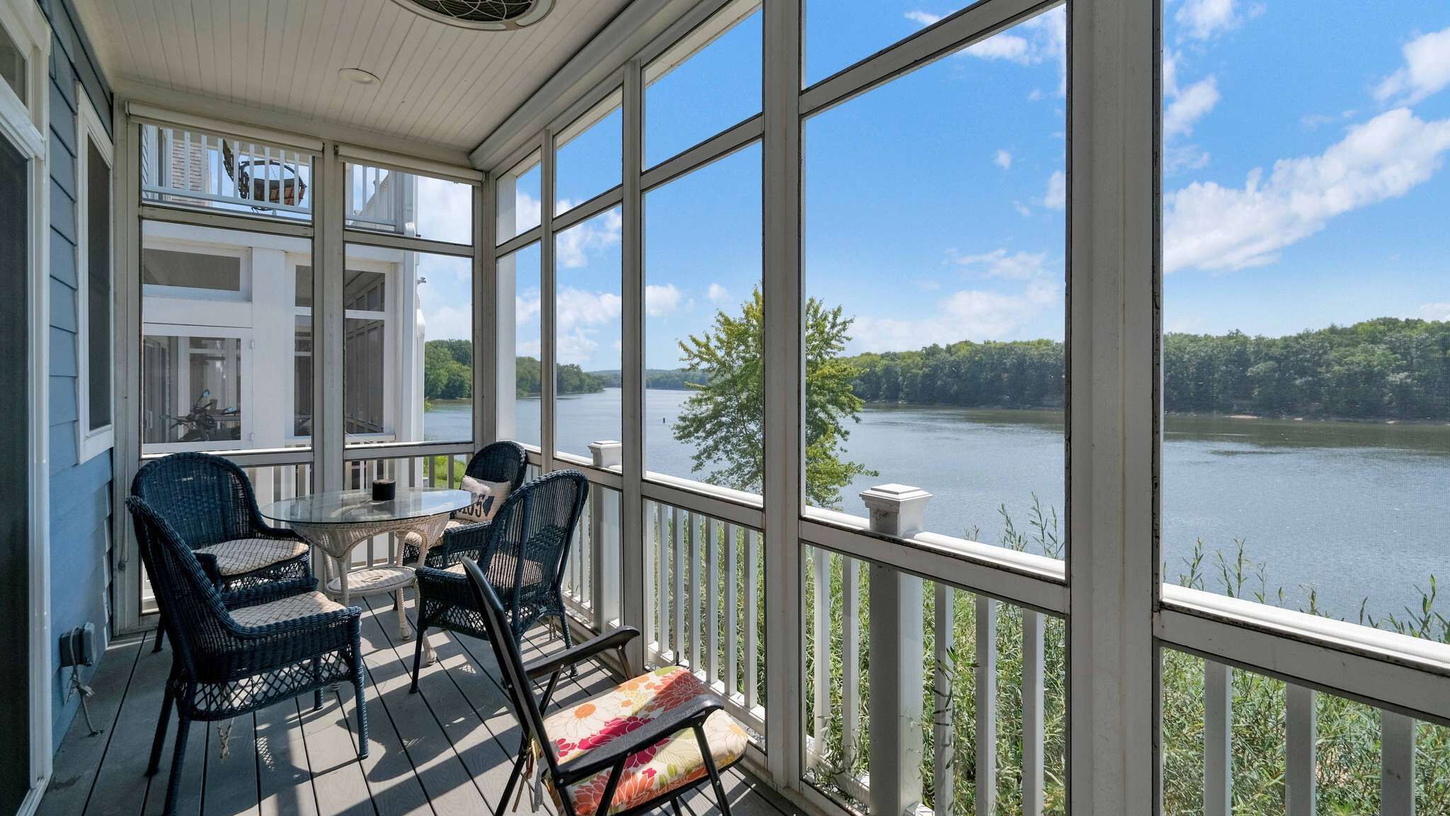Eine überdachte Veranda mit Korbstühlen und einem Tisch, mit Blick auf einen See mit Bäumen und einer Anlegestelle.