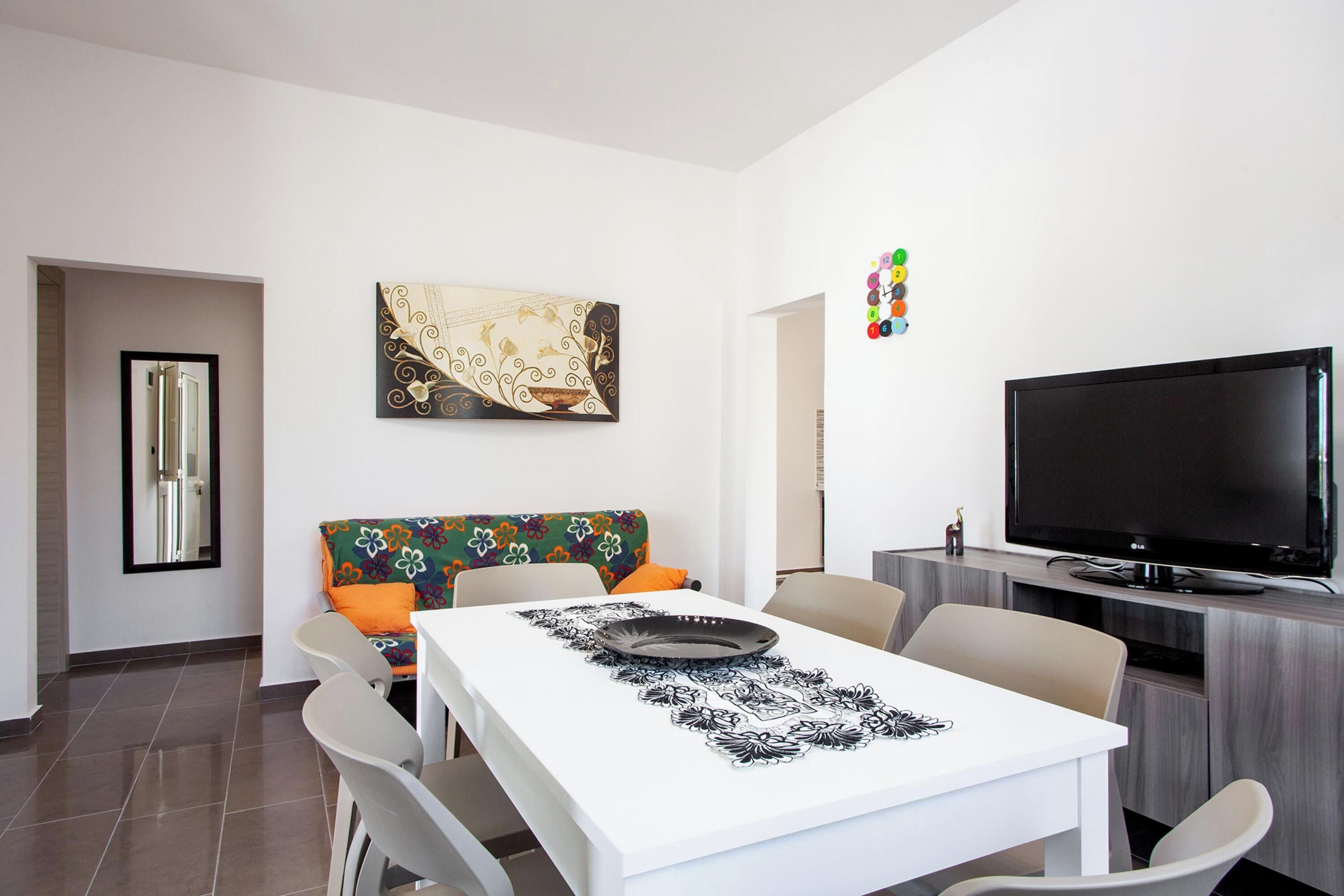 Een moderne woonkamer met een eettafel, stoelen, een bank met kleurrijke kussens, een flatscreen televisie en een decoratief wandobject.